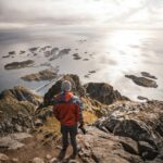 Man explores breathtaking view of Lofoten Islands from mountain peak at sunset.